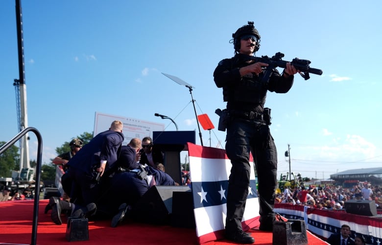 An armed law enforcement officials looks on as a group of security guards surround someone on the ground.