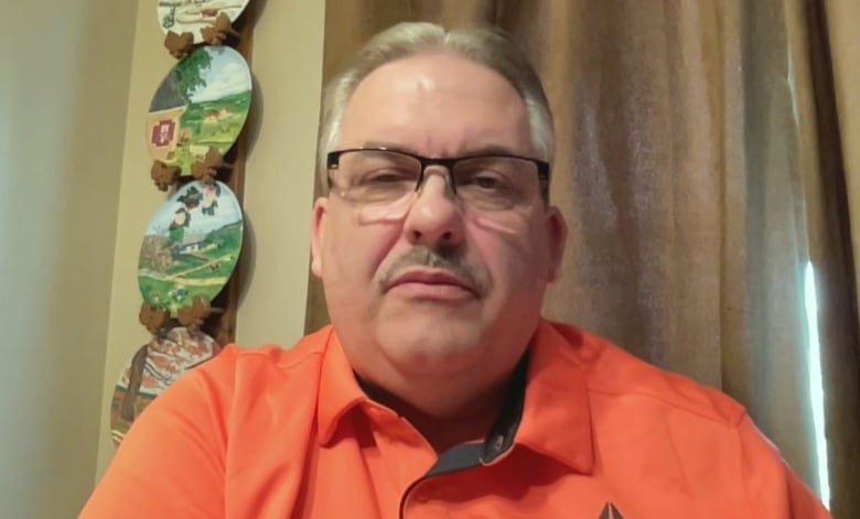 A man in an orange shirt sitting in front of a window with curtains. 