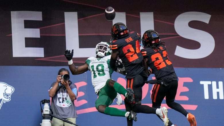 A football player wearing white and green falls while looking at a ball above him, while two others in black follow.