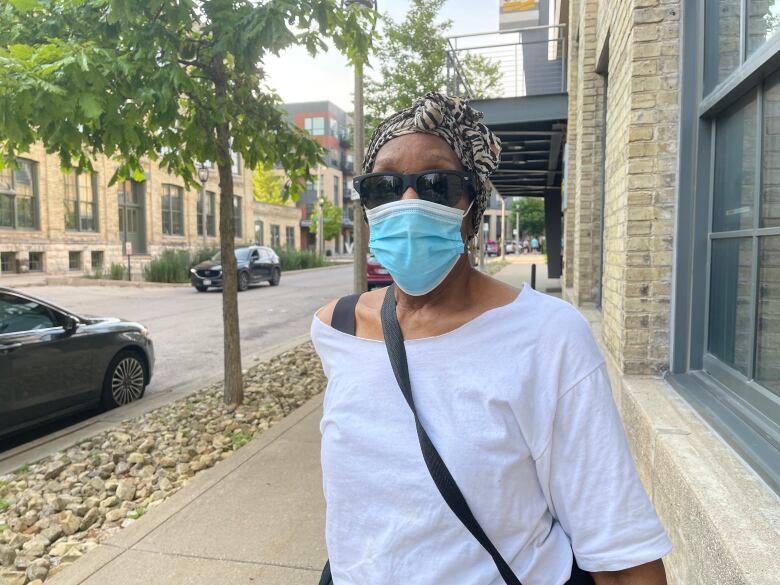 An older woman wearing black sunglasses and a mask is shown on a quiet street.