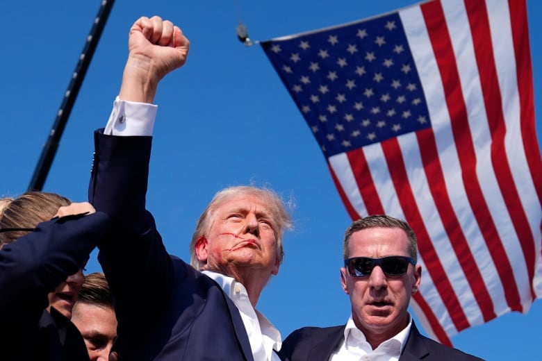 A clean-shaven older man with blood on his face raises his right fist while surrounded by a handful of people who, like him, are wearing dark suit jackets.