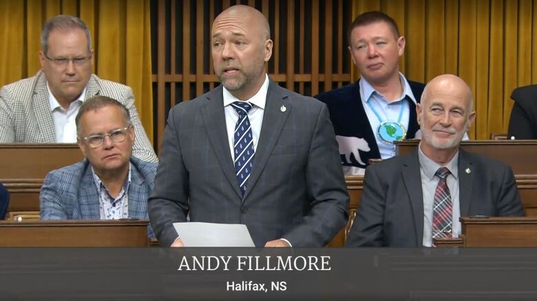 A white, bald man in a grey suit and blue tie satnds among wooden benches.  Four men in suits are seen sitting around him