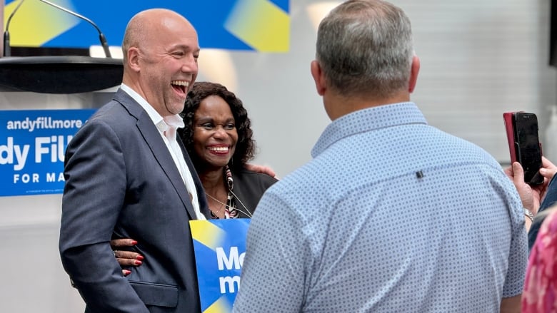 A bald, white man in a navy suit and white shirt laughs with his arm around a smiling Black woman holding a blue sign