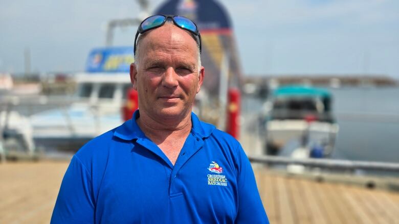 A man with sunglasses perched on his head and wearing a blue shirt stands on a dock in front of a boat.