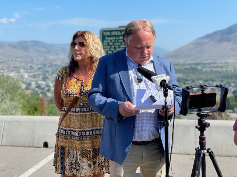 A man speaks to a mic with a woman behind him, with hills visible behind him.