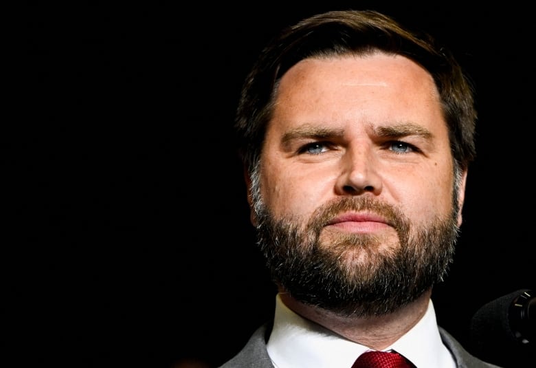 A close-up photo of a man with a full beard wearing a dark suit and red tie.