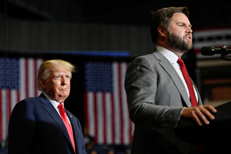 A man in a suit with a red tie watches another man speaking at a podium. A large American flag is in the background.