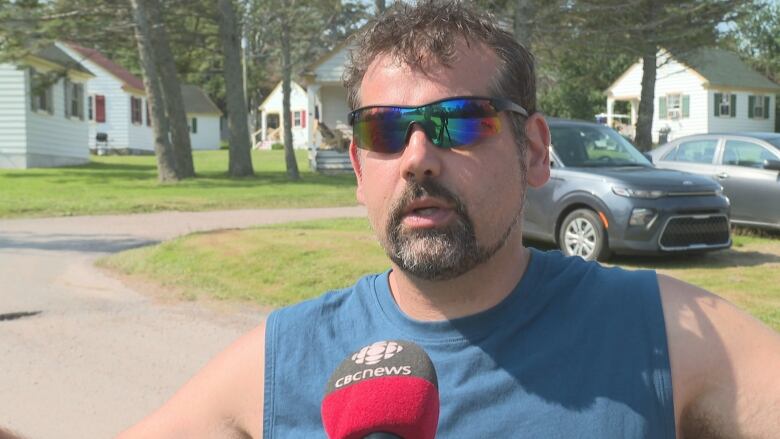 Man in blue sleeveless shirt and sunglasses talks into a microphone.