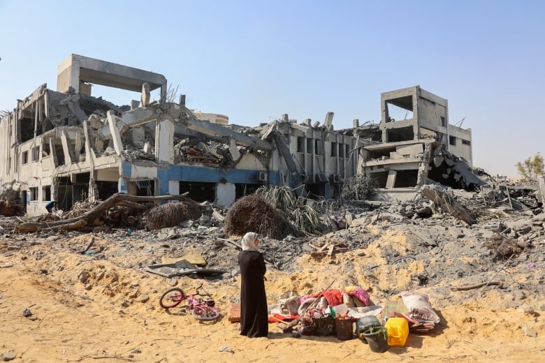 Wide shot of a heavily damaged building. In front of the building is a woman looking on.