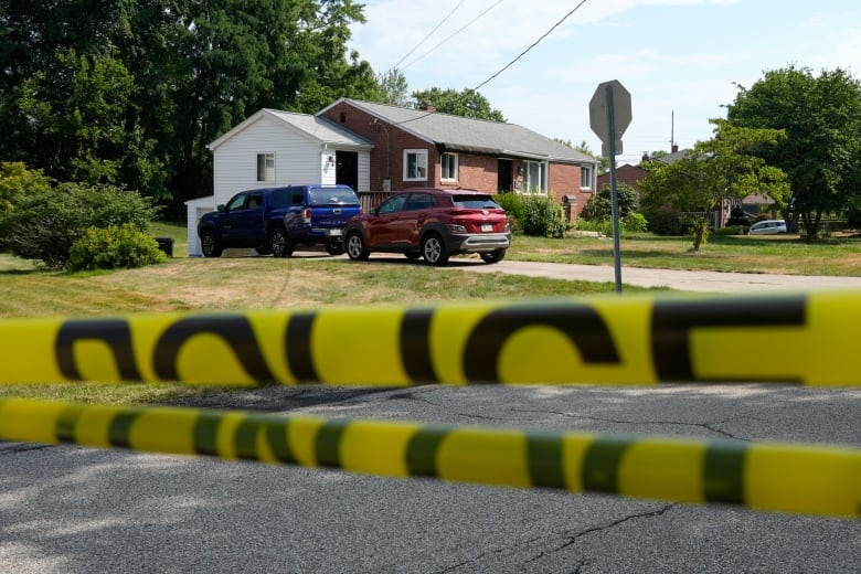 Police tape is shown in the foreground as a one-storey house with two vehicles in the driveway is shown in the background.