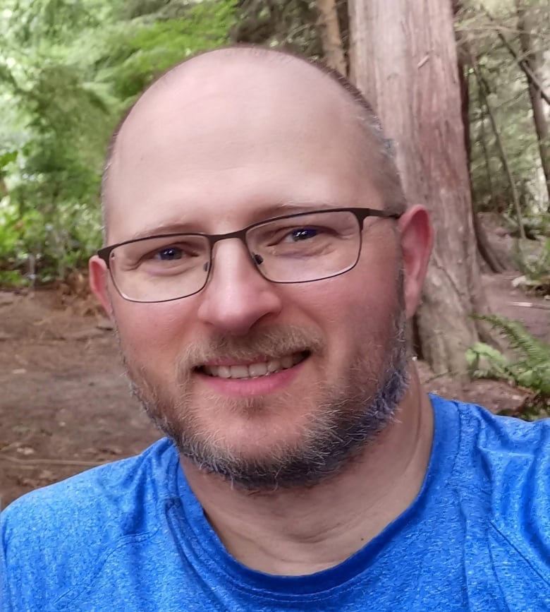 A bald man with a short grey beard and rectangular glasses smiles.