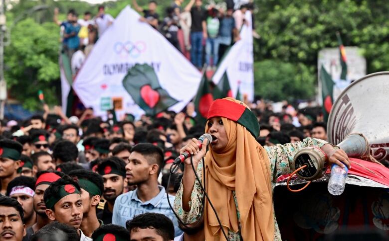 A young woman wearing a covering on her head speaks into a microphone in front of a large outdoor gathering.