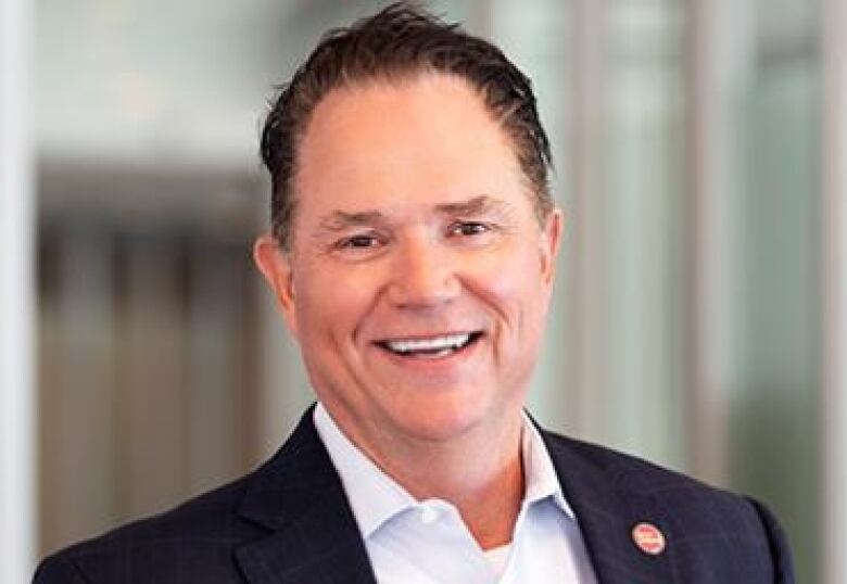 A man smiles for a portrait photograph. He is wearing a suit jacket and collared shirt, but no tie.