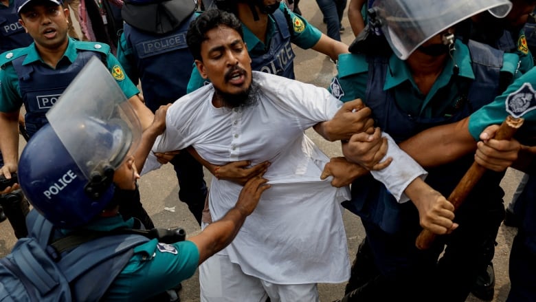 Police in full riot gear wrestle with a man wearing white clothing.