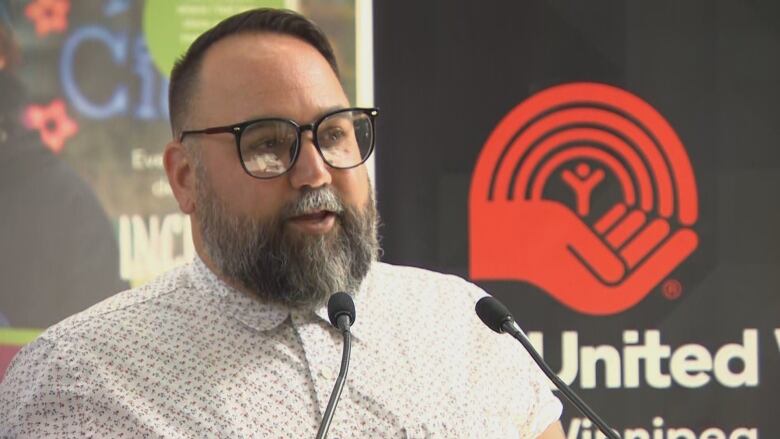 A man in short dark hair and eyeglasses, wearing a button-up collared shirt, speaks into a microphone.