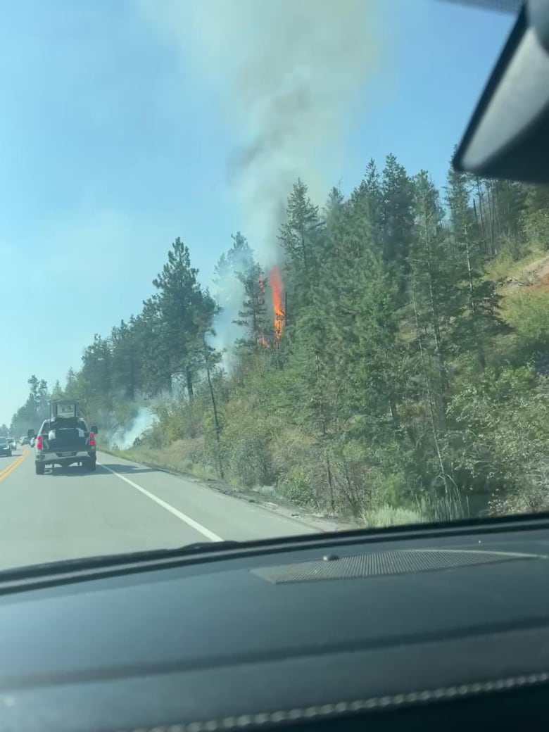 A pickup truck drives by a smoky fire creeping up a hillside next to a highway.