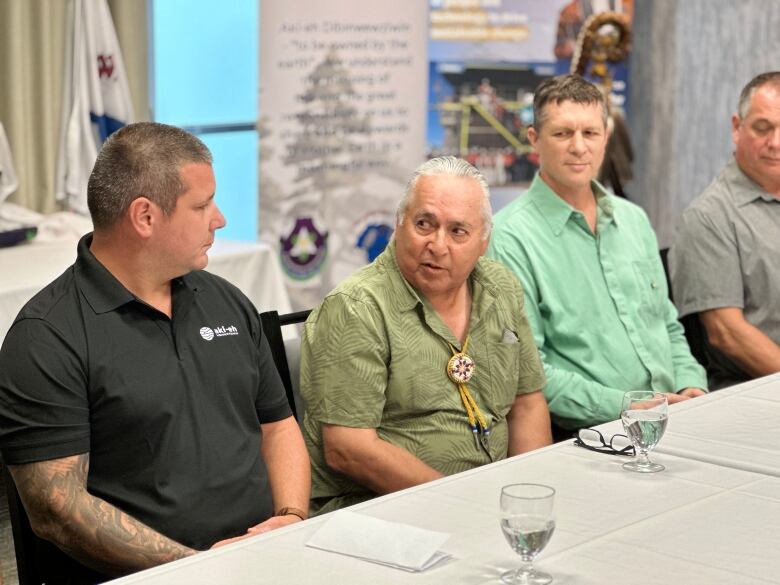 A man with silver hair speaks to another man at a table at a news conference