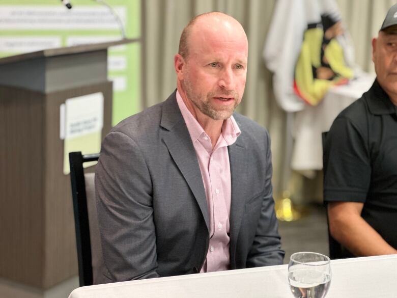 A bald man in a suit speaks at a news conference. He's seated at a table indoors