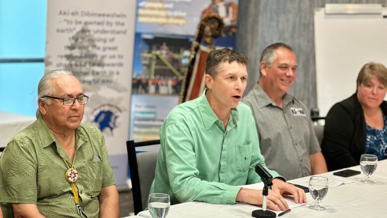 Four people are seated at a table at a news conference. The man in the middle speaks into a microphone as the others listen