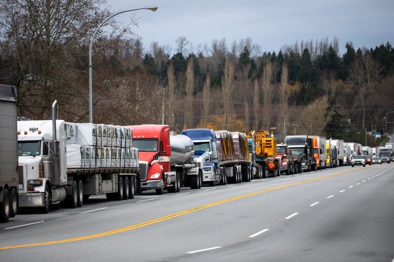 A long line of semi-trucks sit on the side of a highway as cars drive past them in another lane. 