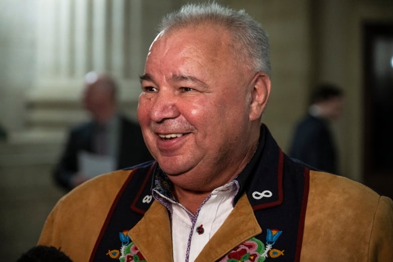 A man in short white hair and a suede jacket smiles and is seen from the shoulders up.