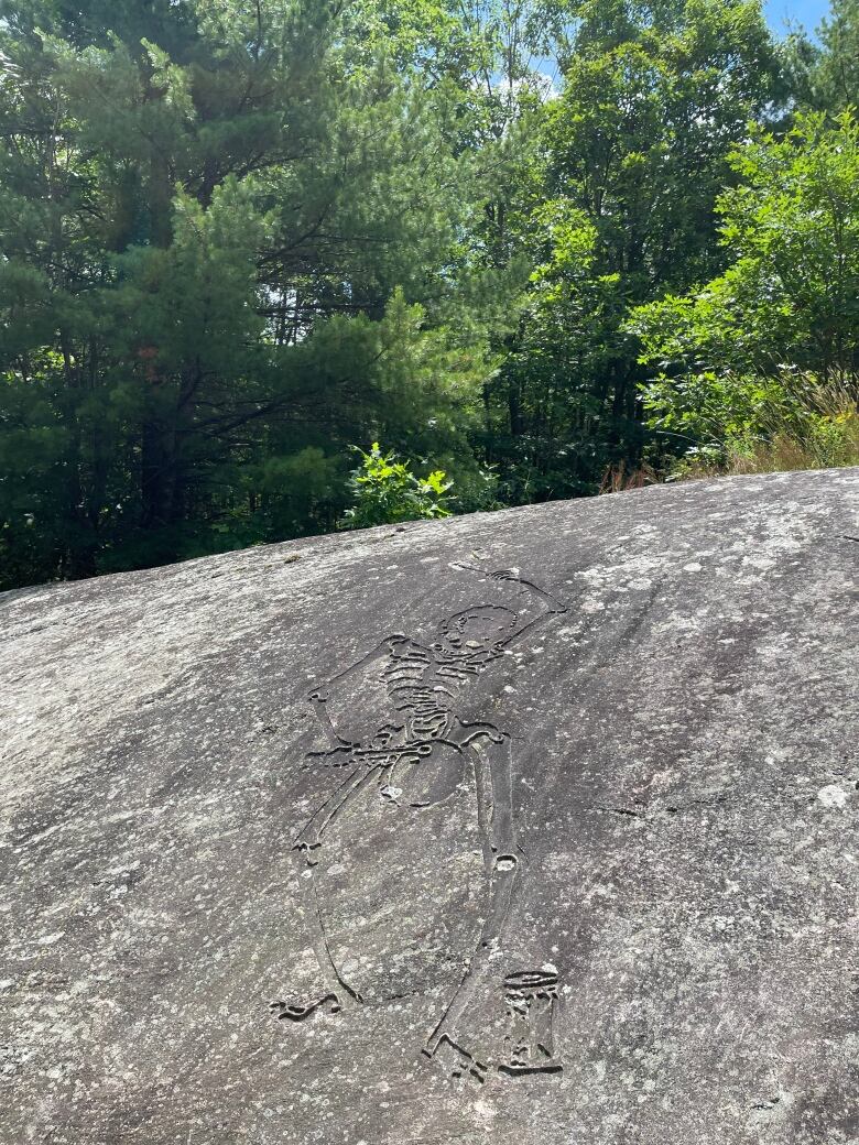 Outdoor scene. An image of a skeleton playing drums has been etched into a giant rock. Green trees rise in the distance.