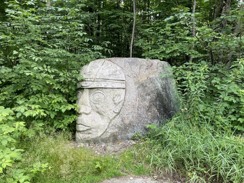 Photo of a large rock carving in the shape of a head, installed in a leafy green woodland field.