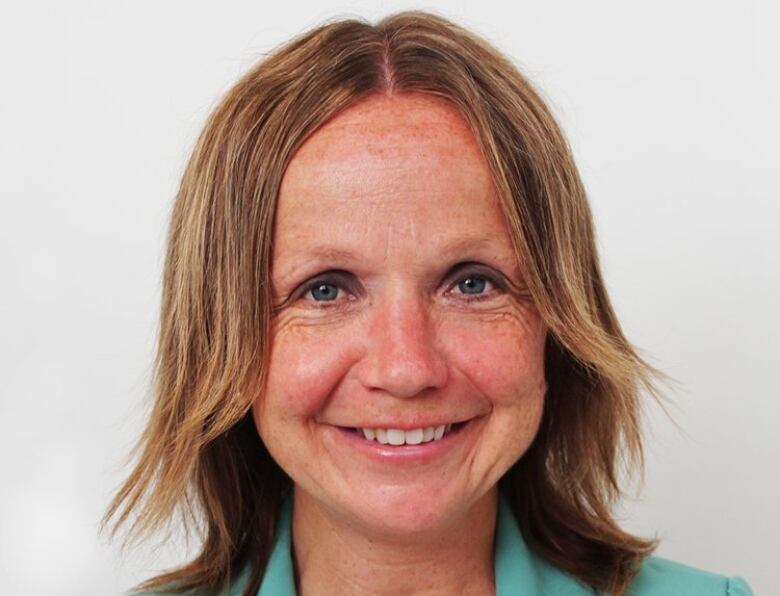 A woman with shoulder-length light brown hair, parted in the middle, and wearing an aquamarine coloured blazer and white shirt smiles at the camera for a head and shoulders portrait against a white background.