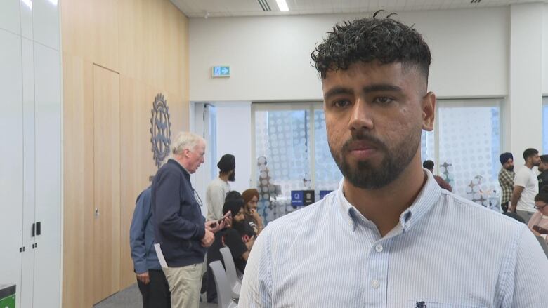 A man looks in to the camera in a blue shirt. In the background are folks chatting to each other after a news conference.