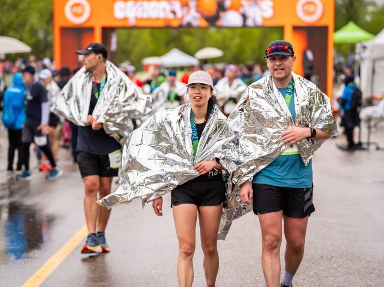 Three runners have just crossed the finish line. They have mylar blankets draped across their shoulders.