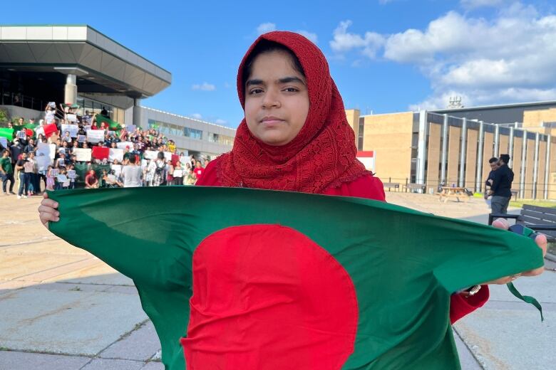 A woman is holding a Bangladesh flag in front of her body. In the background, a large group of people is gathered, holding signs and posters.