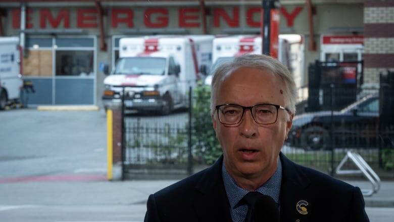 A man with grey hair and glasses stands in front of a hospital. 