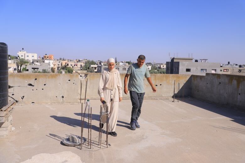 Two people walk on a flat roof of a building, with other buildings in the background.