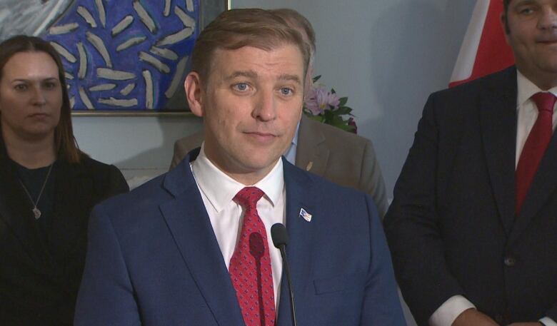 Man in suit stands at mic with people standing behind him