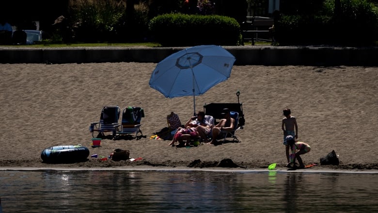 Beachgoers are seen beneath an umbrella, with children playing on the sands.