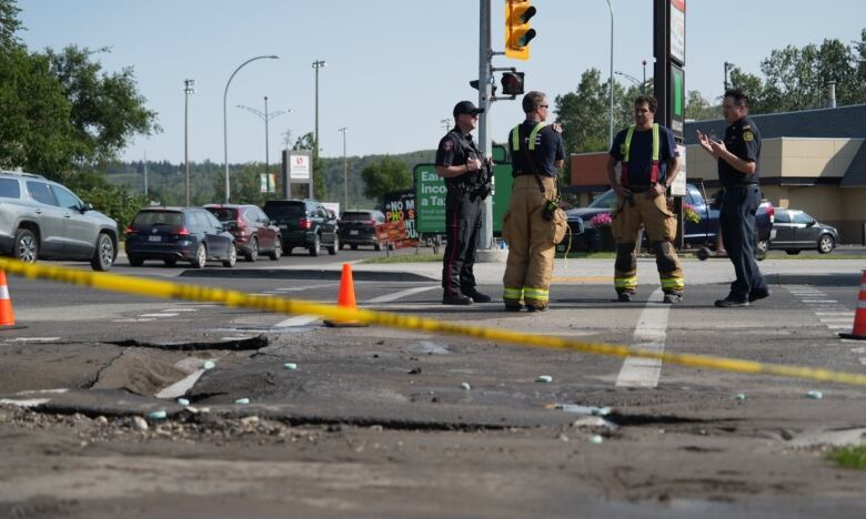 firefighters standing with police officers. there is a broken hole in the road and caution tape around it.