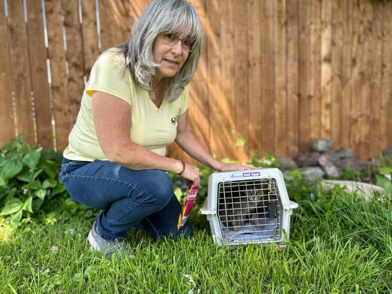 A woman crouches down outdoors next to a small portable cage that has a cat inside.