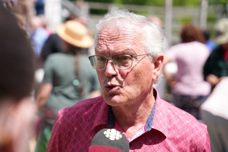 A man with grey hair and glasses speaks to a reporter outdoors on a sunny day.