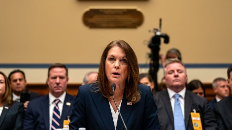 A woman in a blue blazer speaks into a microphone, with a number of people seated behind her. 