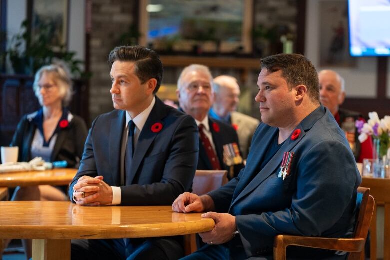 Two men sit at a table in navy suits wearing red poppies. They are looking away from the camera towards something out-of-frame.