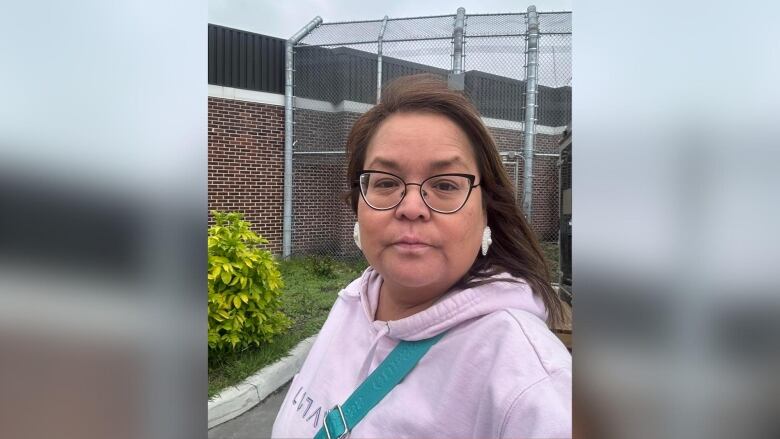 A person wearing a light pink hoodie is seen outside a brick building surrounded by a chain-link fence.