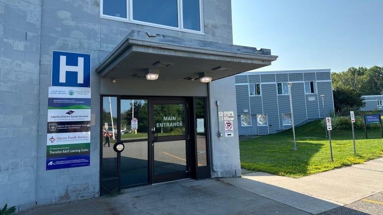 A grey building with a glass door and a sign next to with a large white H on a blue background