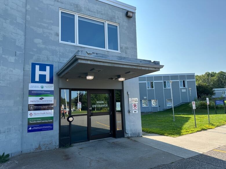 A grey building with a glass door and a sign next to with a large white H on a blue background