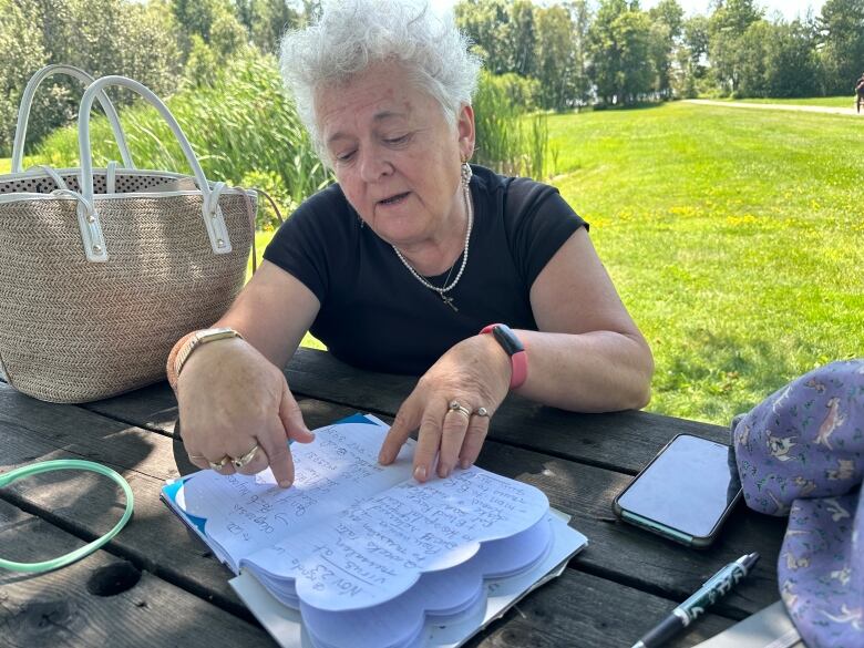 A lady with short grey hair wearing a black t-shirt sits at a picnic table outside. She is pointing to writing in a notebook in front of her.