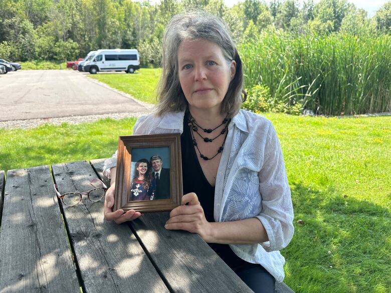 A woman with shoulder length brownish hair and wearing a black bead necklace with a white blouse over a black t-shirt sits at a picnic table holding  a framed portrait of her parents