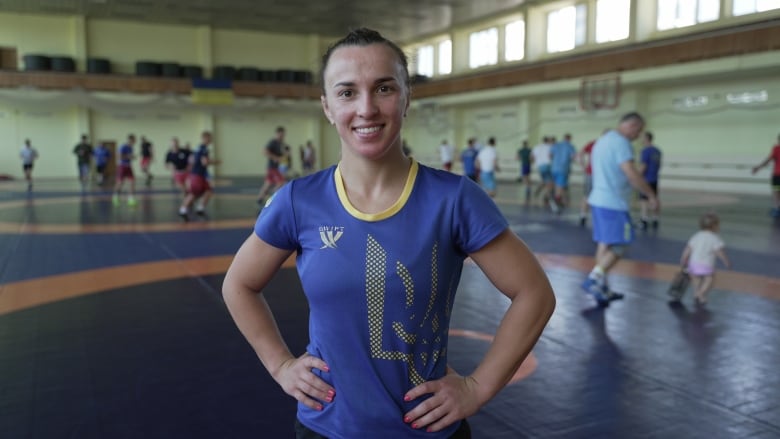 A woman in a blue and yellow shirt stands with hands on her hips in the middle of a gym. 