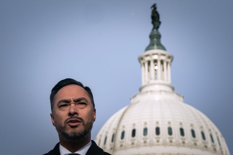 A bearded man is shown speaking with an august building top shown in the background.