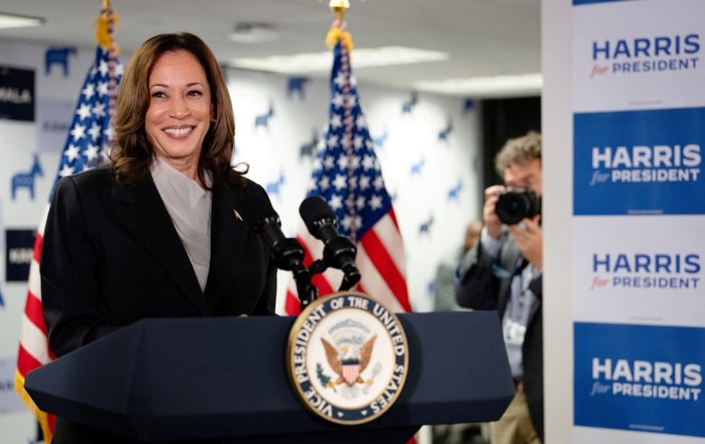 A dark-haired woman in a blazer and blouse smiles as she stands at a podium with an insignia and American flags nearby.