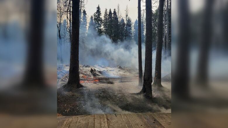 Smoke rises from rubble of a fire in an area surrounded by pine trees.