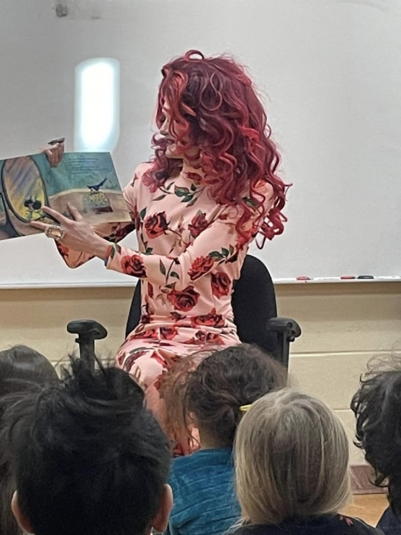 A drag preformer in red wig sitting at an office chair holding a book, children in the foreground.